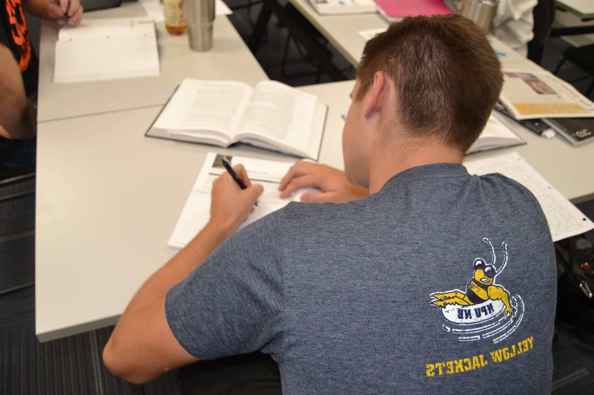 A student wearing a grey shirt with a "Howard Payne University Yellow Jackets" emblem is writing notes in a classroom setting. | HPU